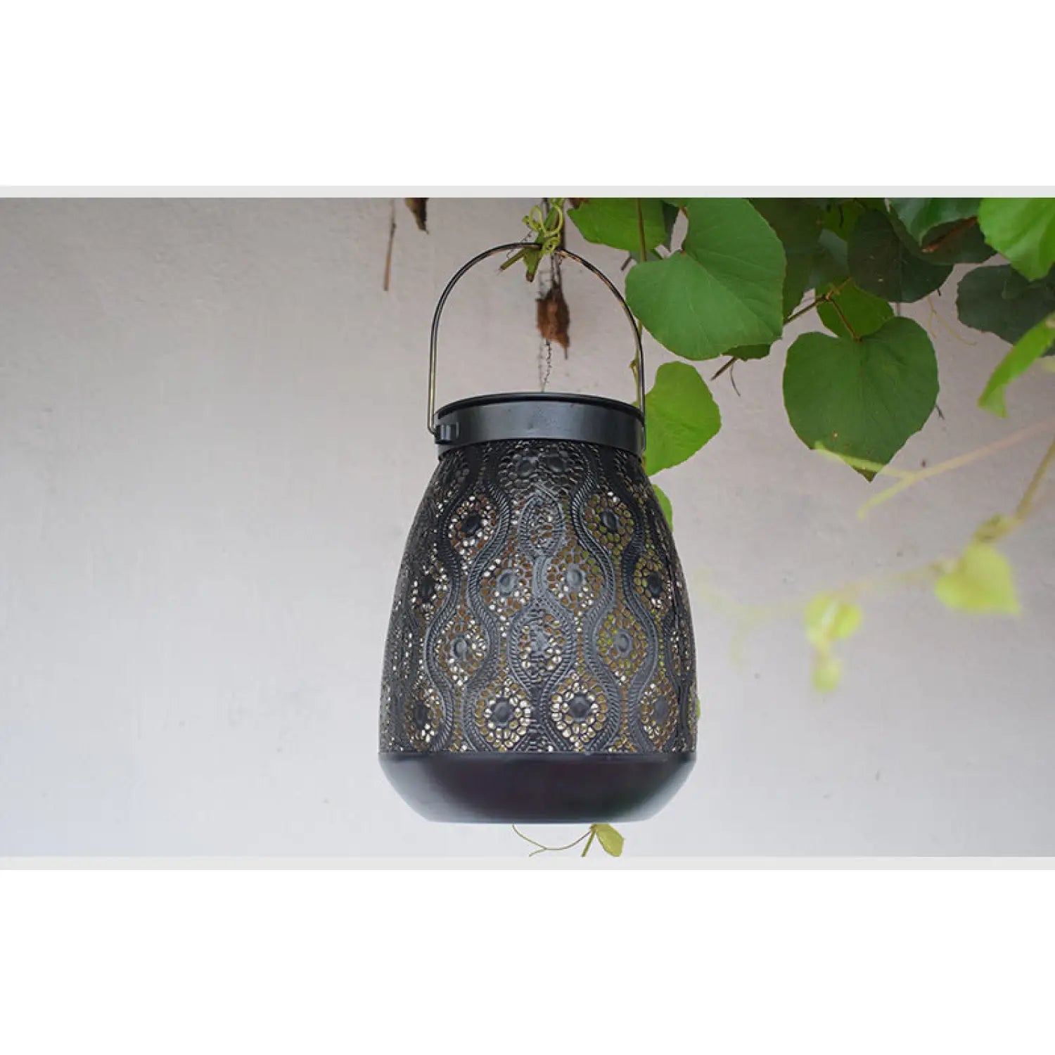 Decorative black lantern with intricate patterns hanging against a light background with green leaves.